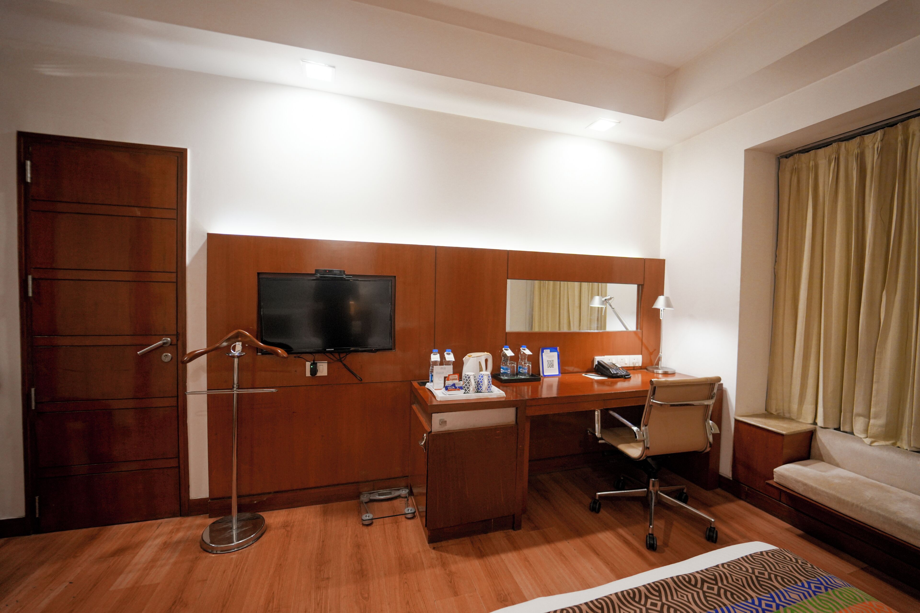 A hotel room with a wooden desk, a television, a bed, a desk lamp, and a small cabinet.