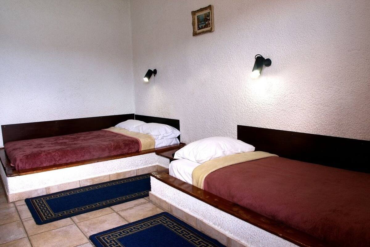 A room with two beds, each with a wooden headboard and a red blanket. There are two wall-mounted lights and a small framed picture on the wall.