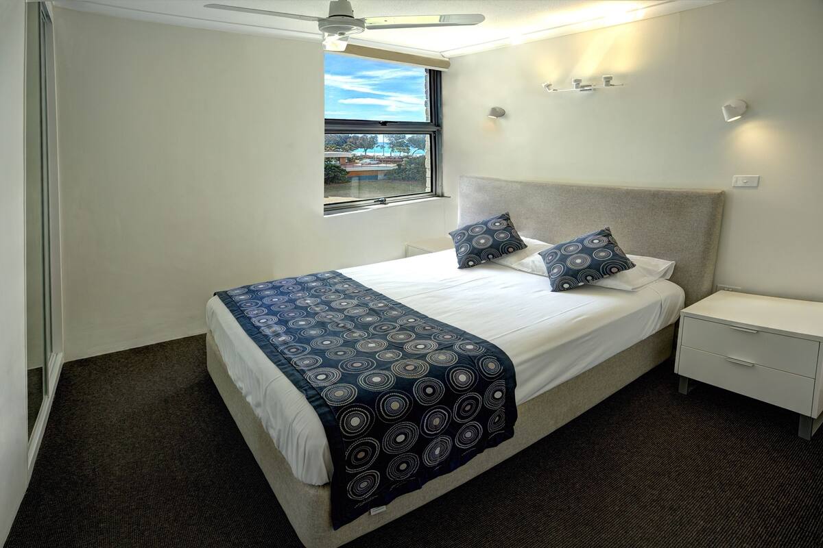 A bedroom with a bed, bedside table, and a window with a view of buildings.