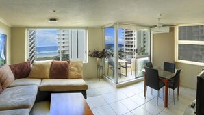 Uma sala de estar moderna com sofá, mesa de jantar com cadeiras e varanda com vista para o oceano e edifícios.