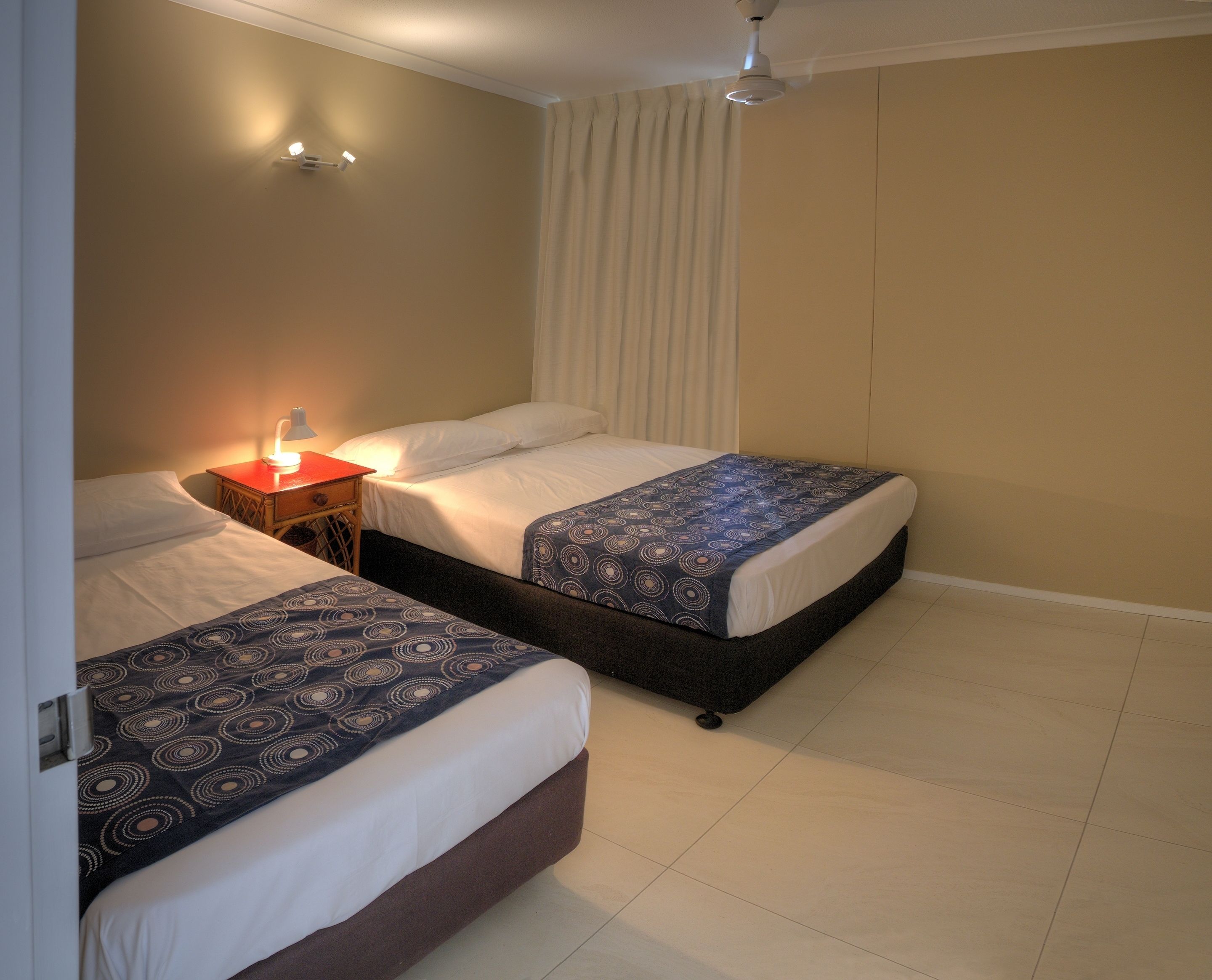 Une chambre d’hôtel avec deux lits, une table de chevet avec une lampe, un ventilateur de plafond et une fenêtre avec des rideaux.