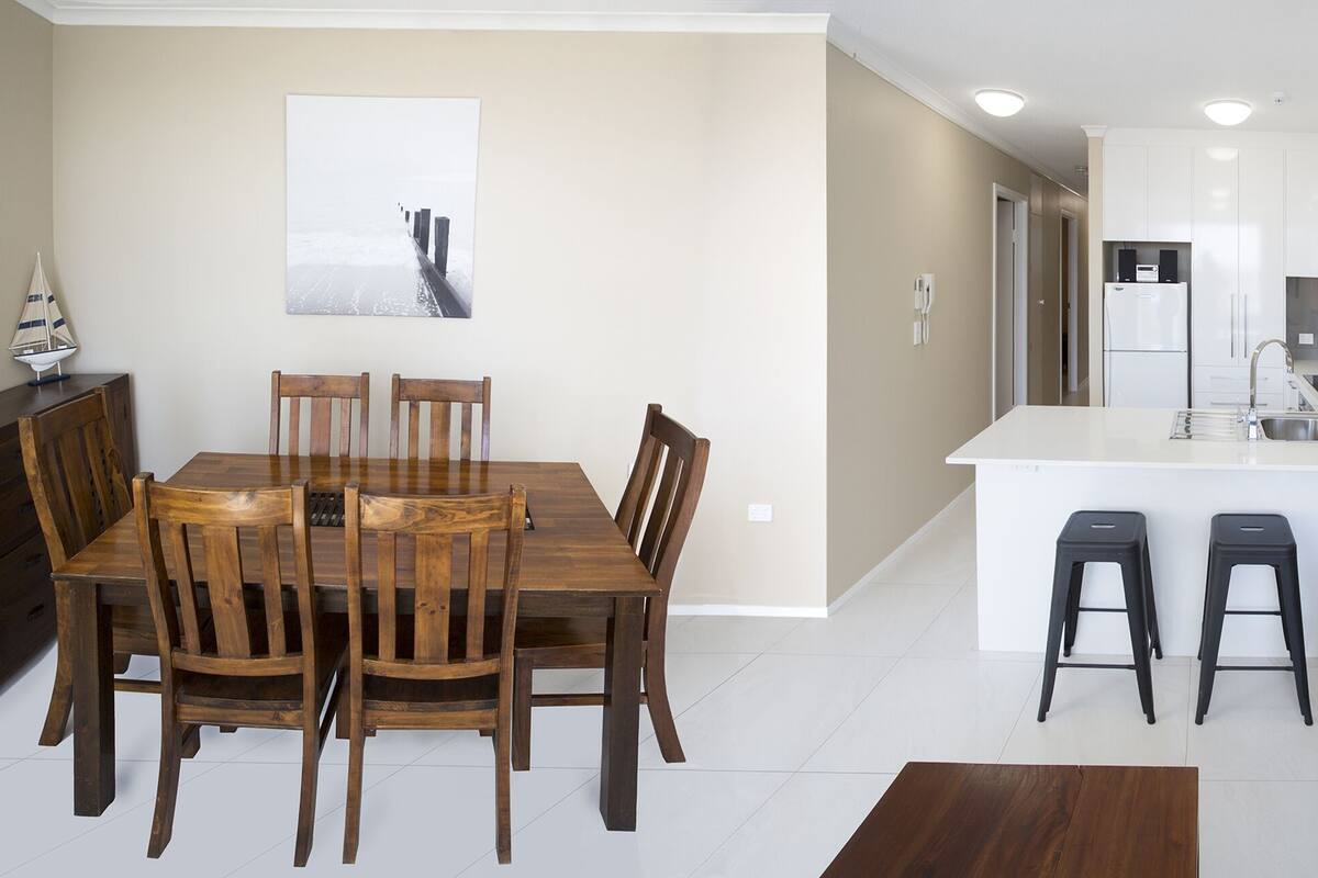 A modern kitchen with white cabinets, a central island with stools, and a dining area with wooden chairs.