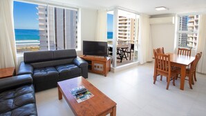 Sala de estar com sofá de couro preto, mesa de centro de madeira e suporte para TV. Há uma área de jantar com mesa e cadeiras de madeira. Janelas amplas oferecem vista para o oceano e prédios.