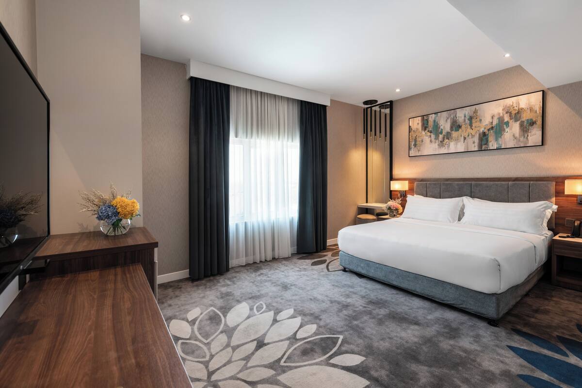 A modern hotel room with a large bed, a desk, a chair, a TV, and a window with curtains.