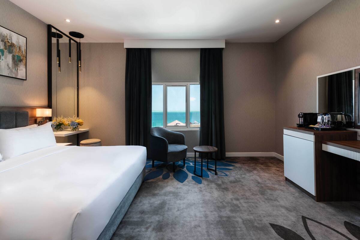 A hotel room with a bed, a desk, a chair, a small table, and a view of the sea.