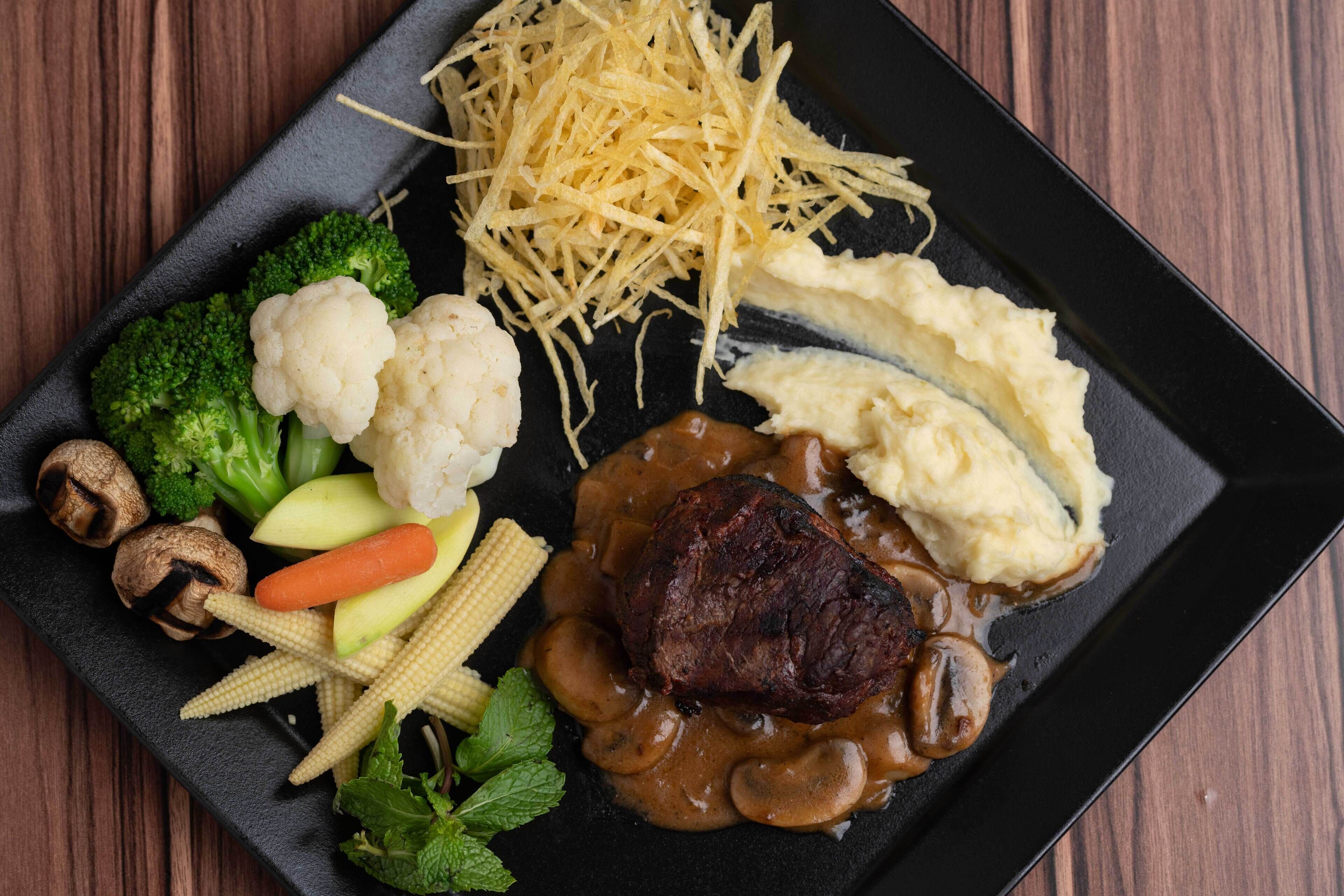A black plate with a steak, mashed potatoes, and a mushroom sauce.