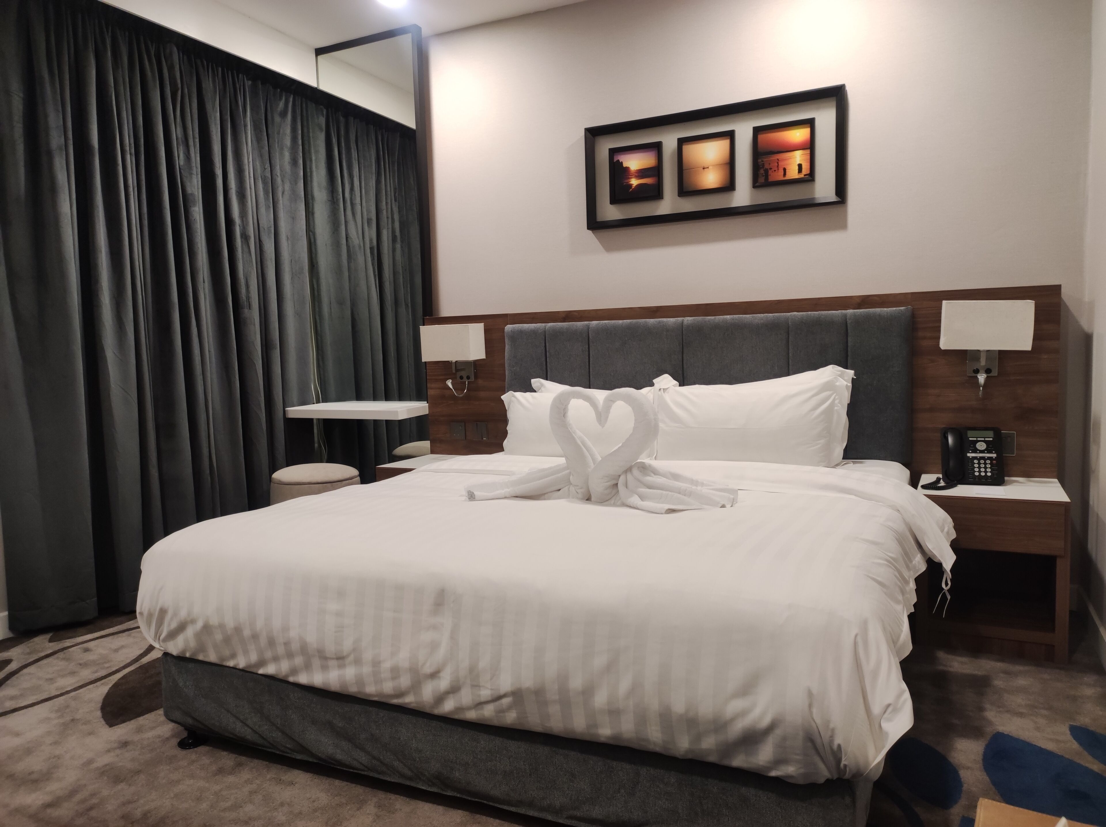 A neatly made bed with white linens, a heart-shaped towel arrangement, a bedside table with a lamp, a phone, and a wall-mounted picture frame.