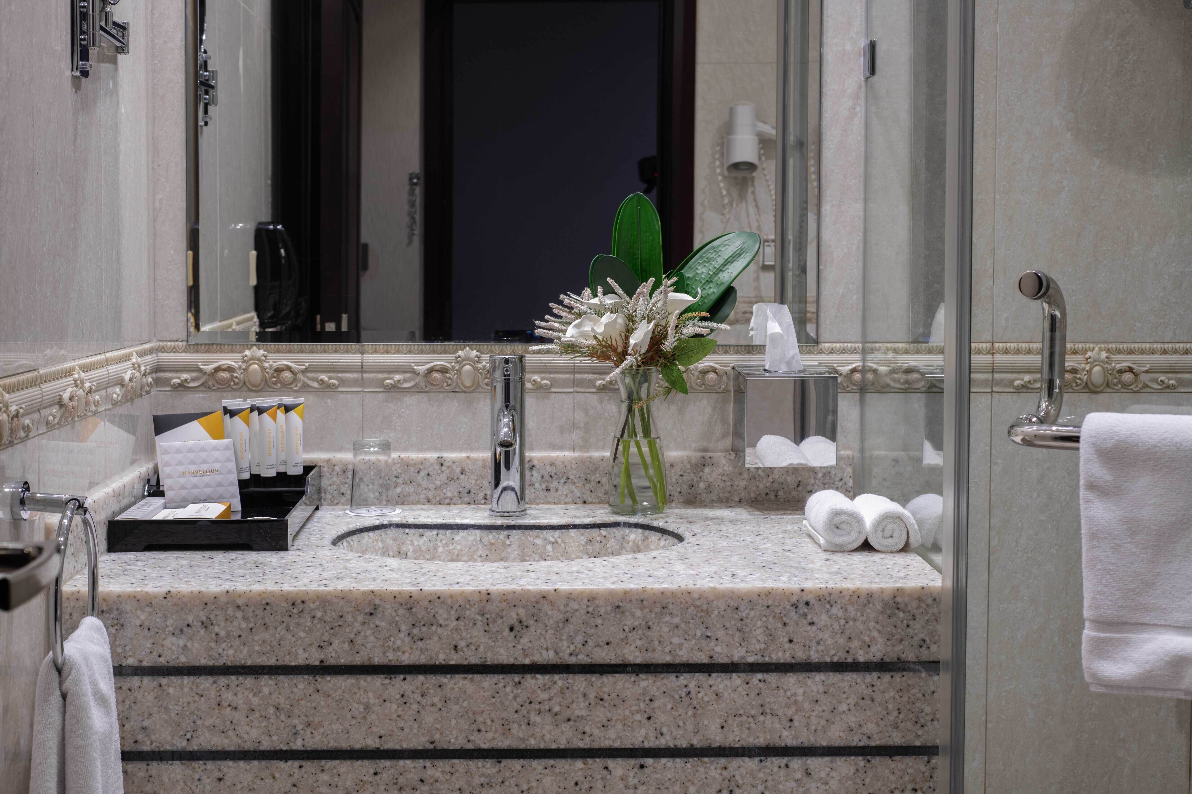 Un baño con encimera de granito, lavamanos, espejo, un florero con flores y un perchero para toallas.