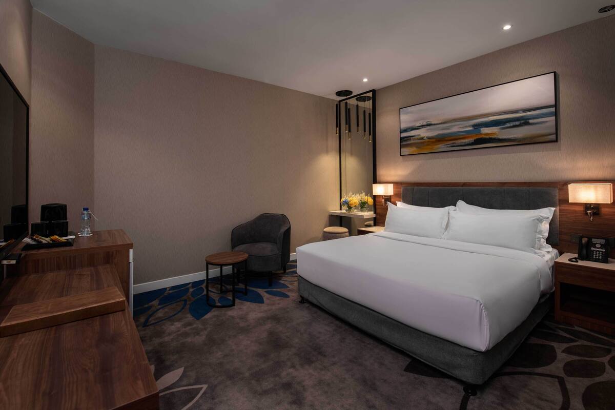 A hotel room with a large bed, a desk, a chair, a small table, and a nightstand with a lamp.