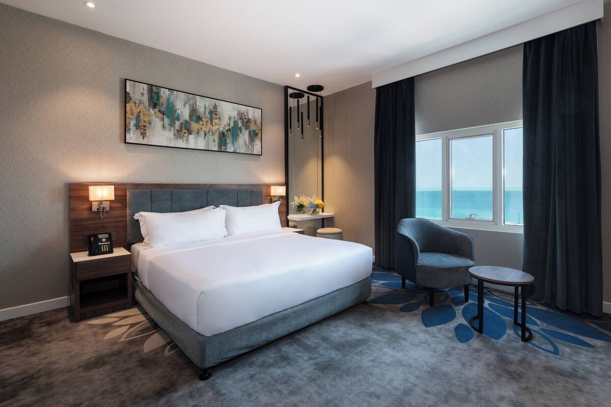 A hotel room with a large bed, a bedside table with a phone, a chair, a small table, a nightstand, a painting on the wall, and a window with curtains.