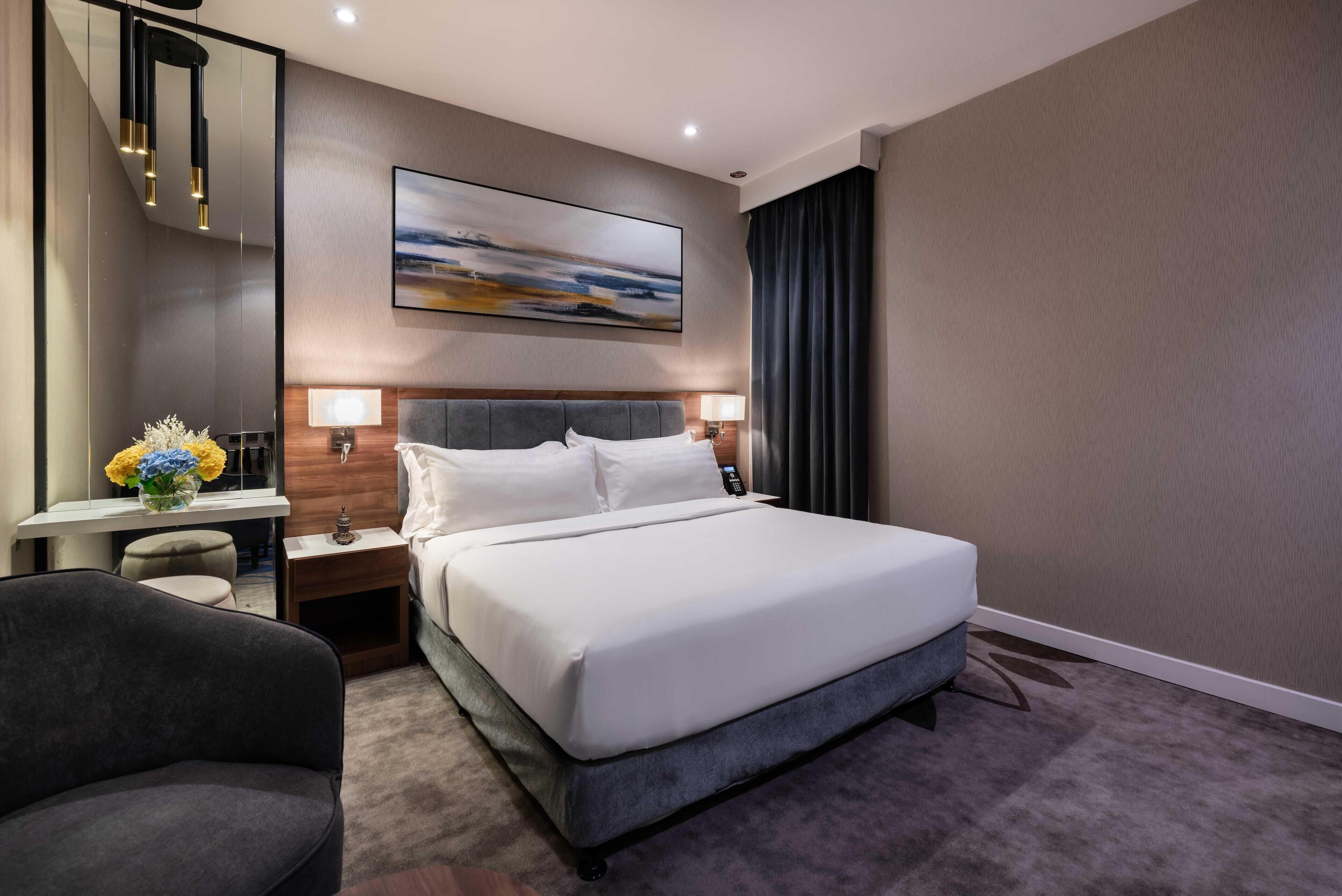 A hotel room with a large bed, bedside tables, a desk, a chair, a painting on the wall, and a window with curtains.