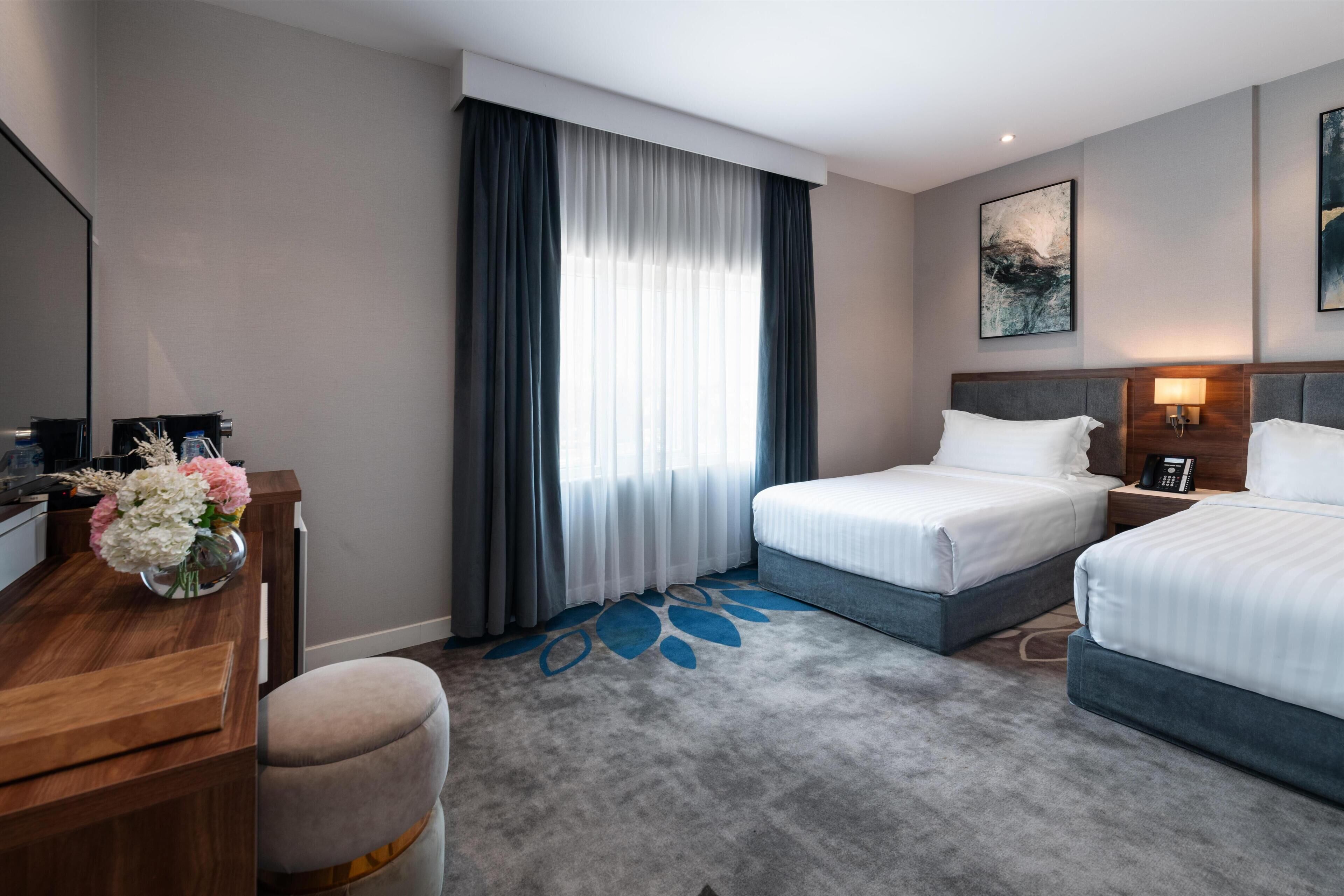 Une chambre d’hôtel avec deux lits, un bureau agrémenté d’un vase de fleurs, une grande fenêtre avec des rideaux et un tableau au mur.