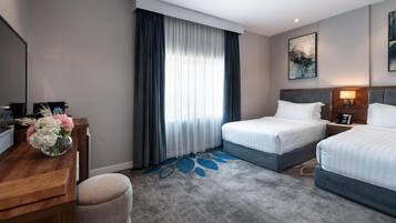A hotel room with two beds, a desk with a vase of flowers, a large window with curtains, and a painting on the wall.