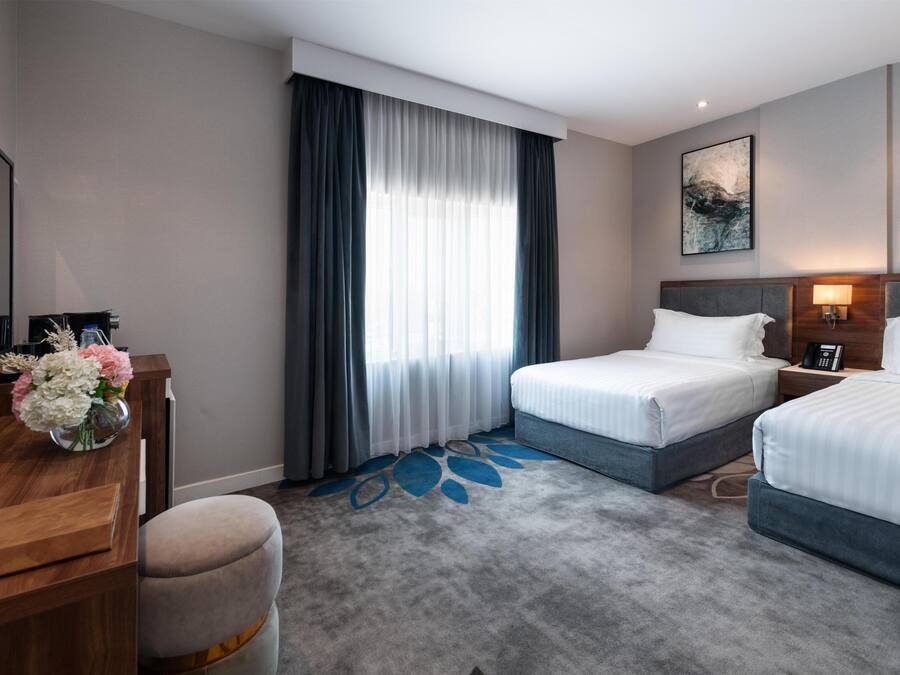 A hotel room with two beds, a desk with a vase of flowers, a large window with curtains, and a painting on the wall.