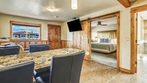 Ein Essbereich mit großem Tisch und Stühlen, einem an einer Holzwand befestigten Fernseher und Blick in ein Schlafzimmer durch eine offene Tür.