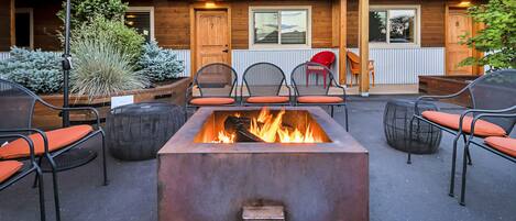 Ein Feuerkorb, umgeben von Sitzgelegenheiten mit Stühlen und einem Tisch, ein Holzgebäude mit Veranda und ein roter Sonnenschirm.