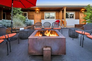 A fire pit surrounded by outdoor seating with chairs and a table, a wooden building with a porch, and a red umbrella.