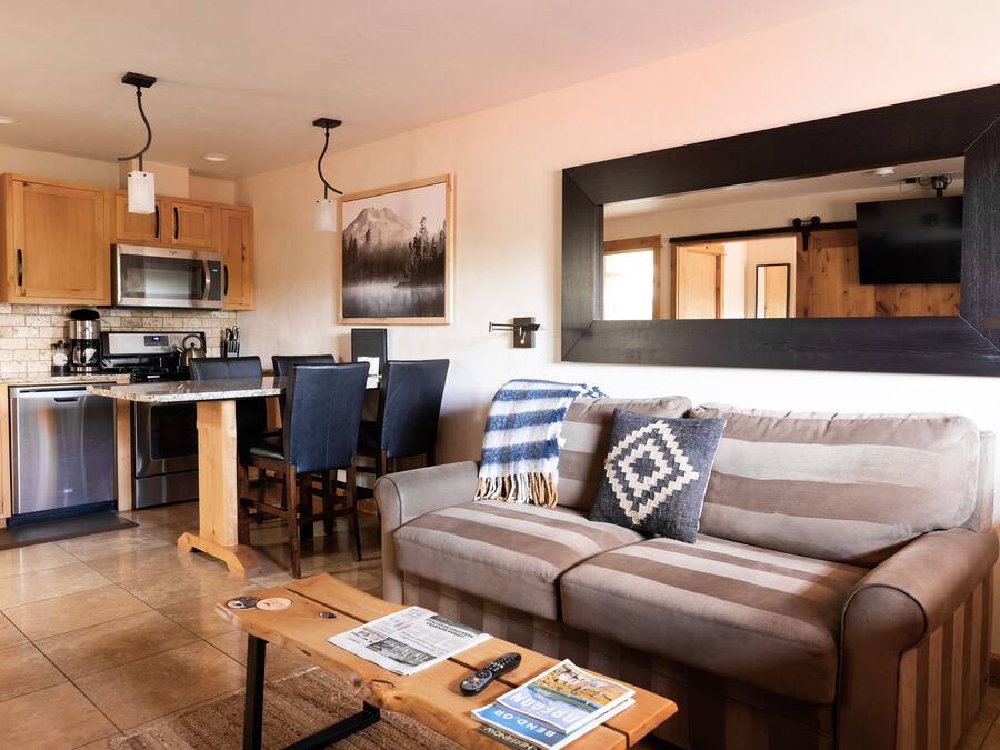 Eine moderne Küche mit Holzausstattung, einer Edelstahl-Mikrowelle und einer Kaffeemaschine. Es gibt einen Essbereich mit Tisch und Stühlen sowie ein Wohnzimmer mit einer Couch und einem großen Spiegel.