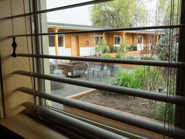 Une vue à travers les stores d’une fenêtre, donnant sur un espace extérieur aménagé en terrasse, avec un bâtiment en bois, du mobilier d’extérieur et de la verdure.