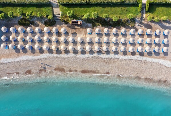 An aerial view of a beach with rows of white umbrellas and lounge chairs, a pathway leading to a building, and clear turquoise water.