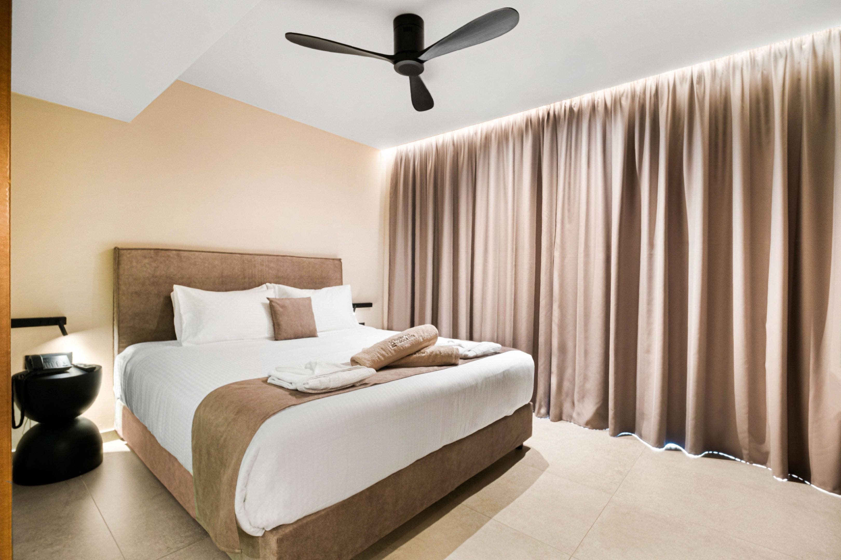A modern hotel room with a large bed, a ceiling fan, and a view of the city through sheer curtains.
