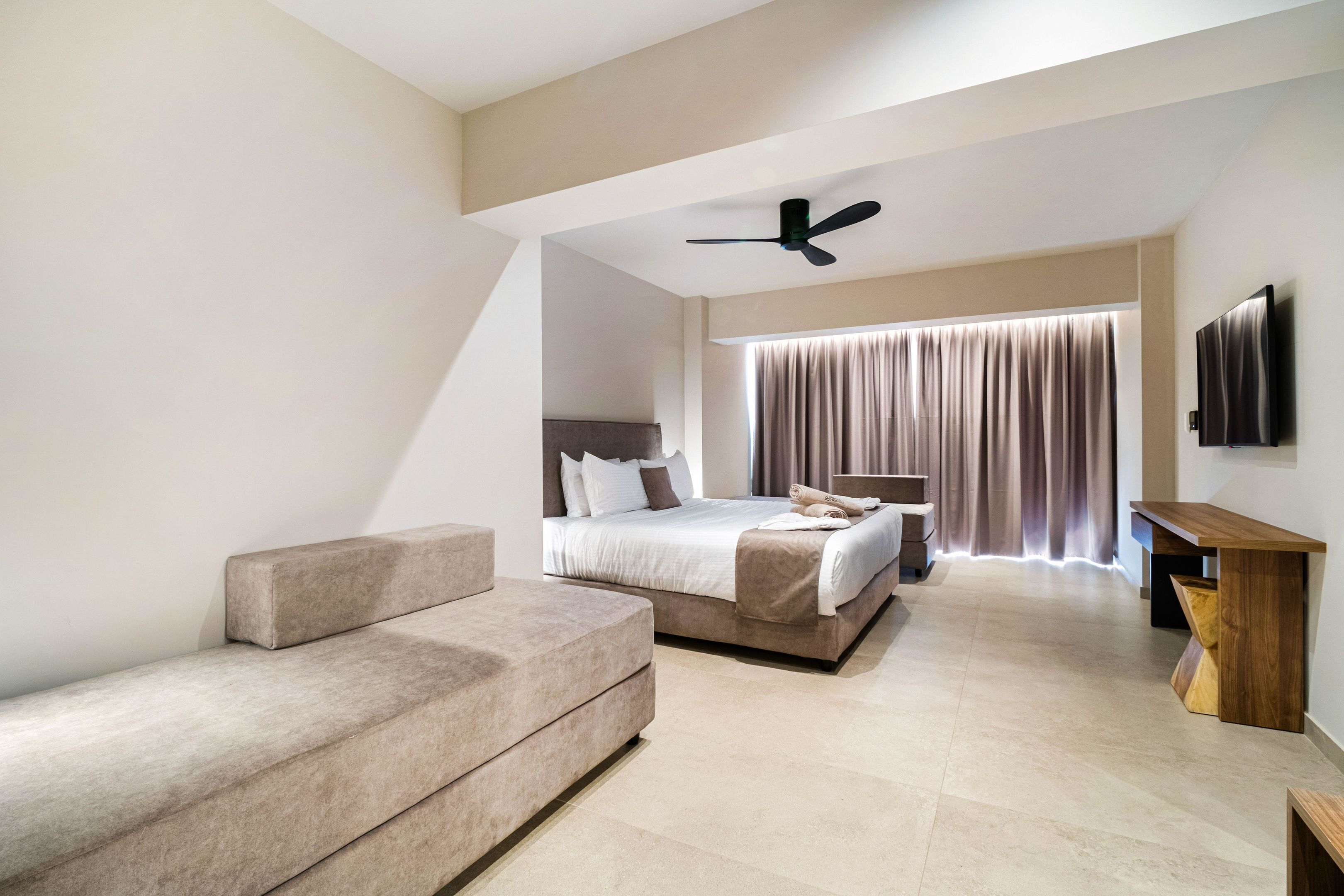 A modern hotel room with a bed, a sofa, a wooden side table, a ceiling fan, and a TV mounted on the wall.