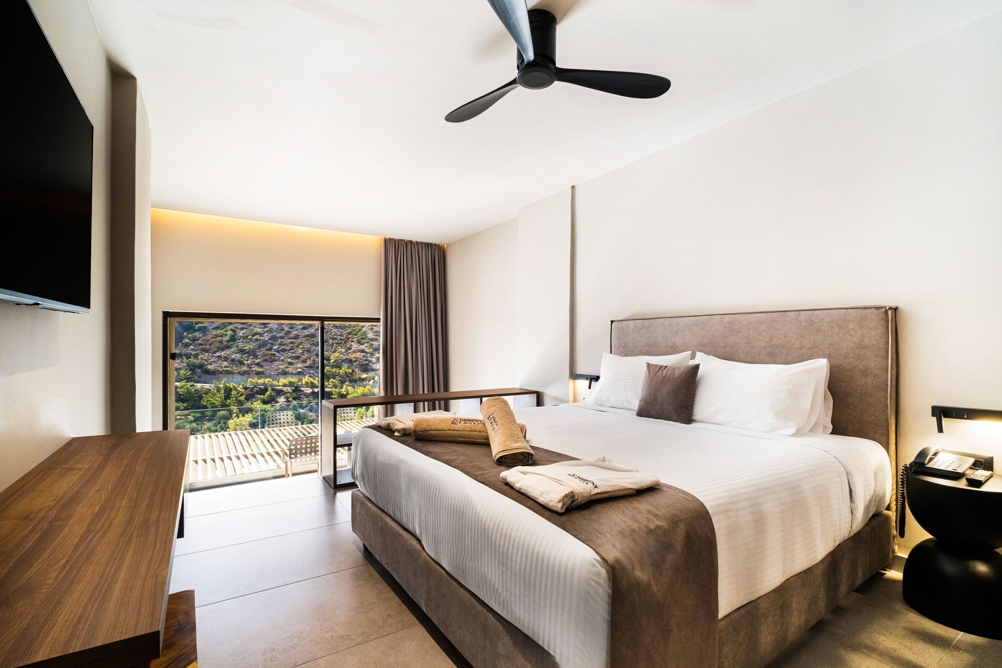A modern hotel room with a large bed, a desk, and a balcony with a view of the hills.