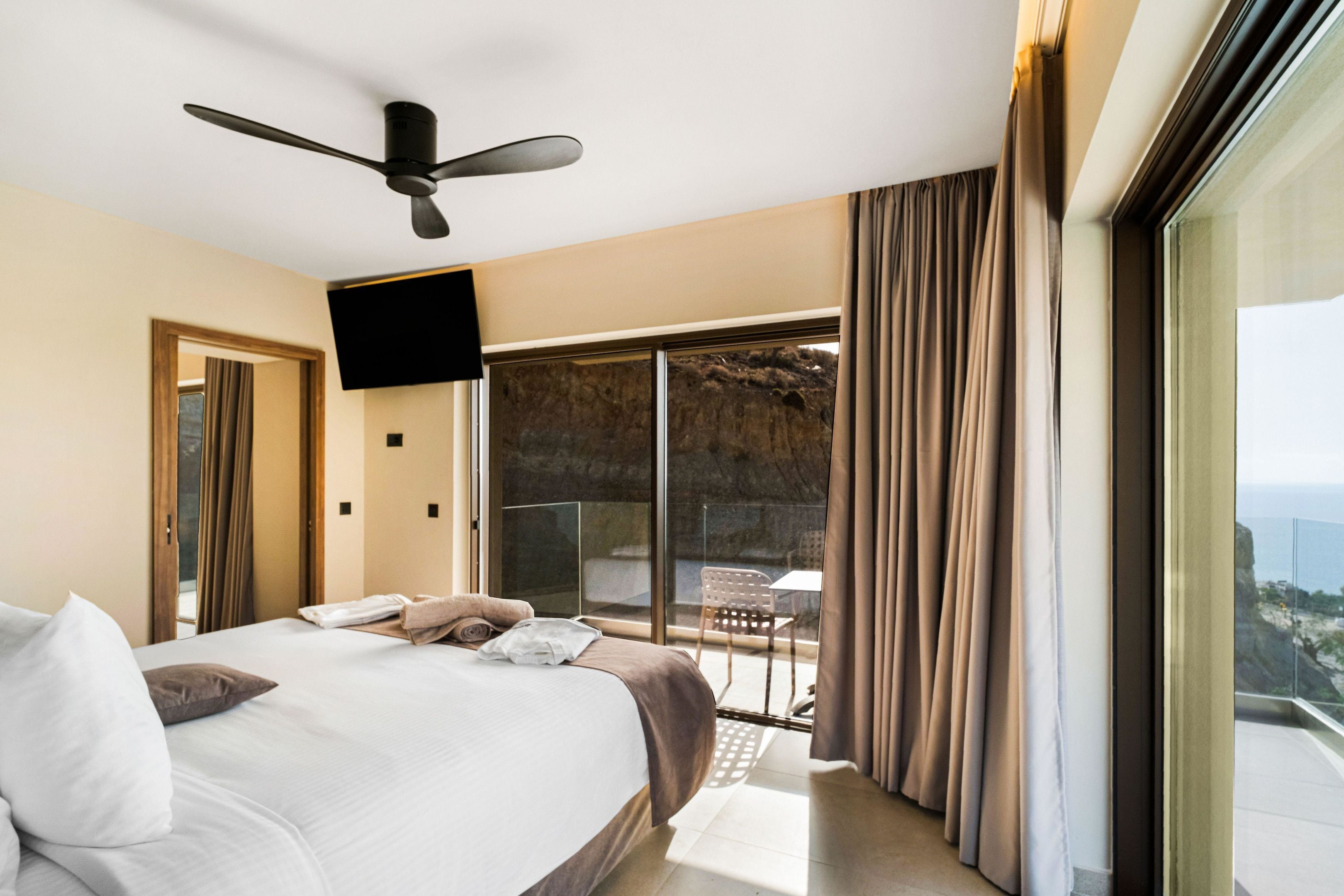 Habitación de hotel con una cama grande, ventilador de techo, televisor y balcón con cortinas.
