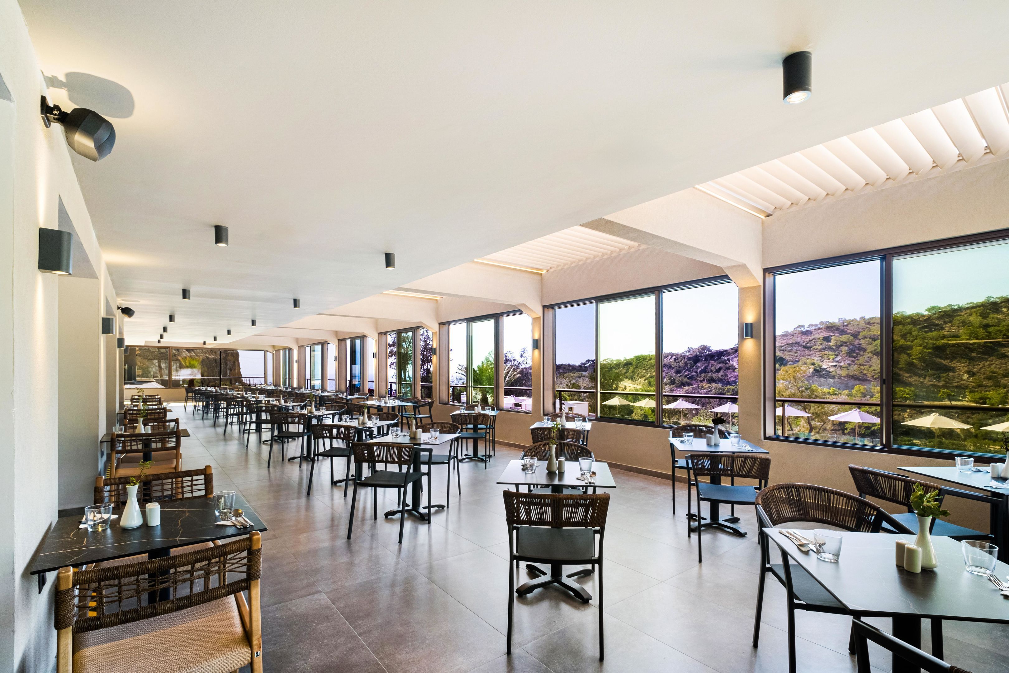 A spacious dining area with high ceilings and large windows offering a view of the outdoors. Tables are neatly arranged with chairs, and the room has a modern, clean design.