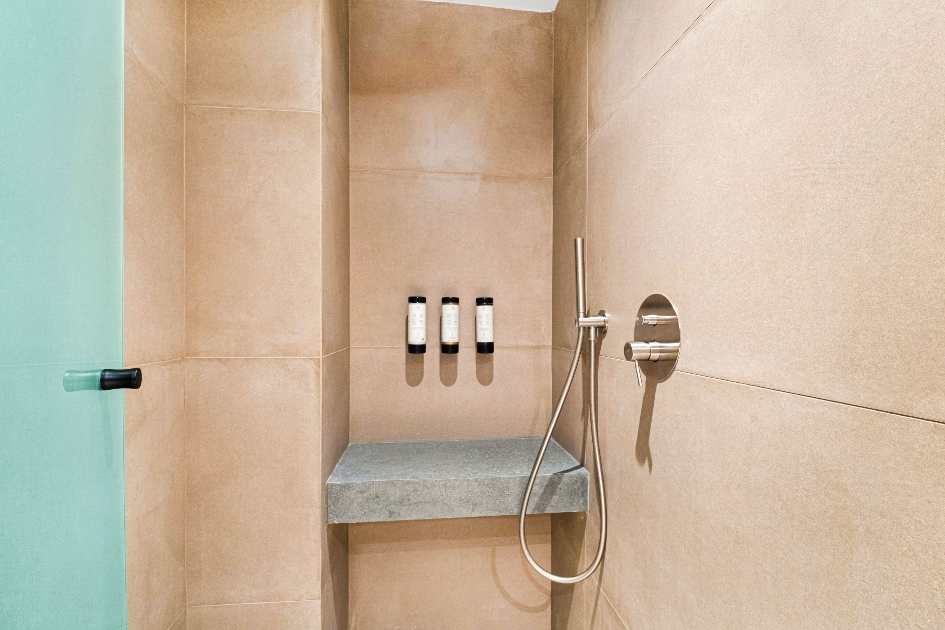 A bathroom with beige tiles, a shower with a handheld showerhead, and three bottles on a shelf.