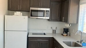 A compact kitchen with dark cabinets, a white refrigerator, a microwave, a sink, and patterned floor tiles. - Oasis Inn & Suites (Santa Barbara)