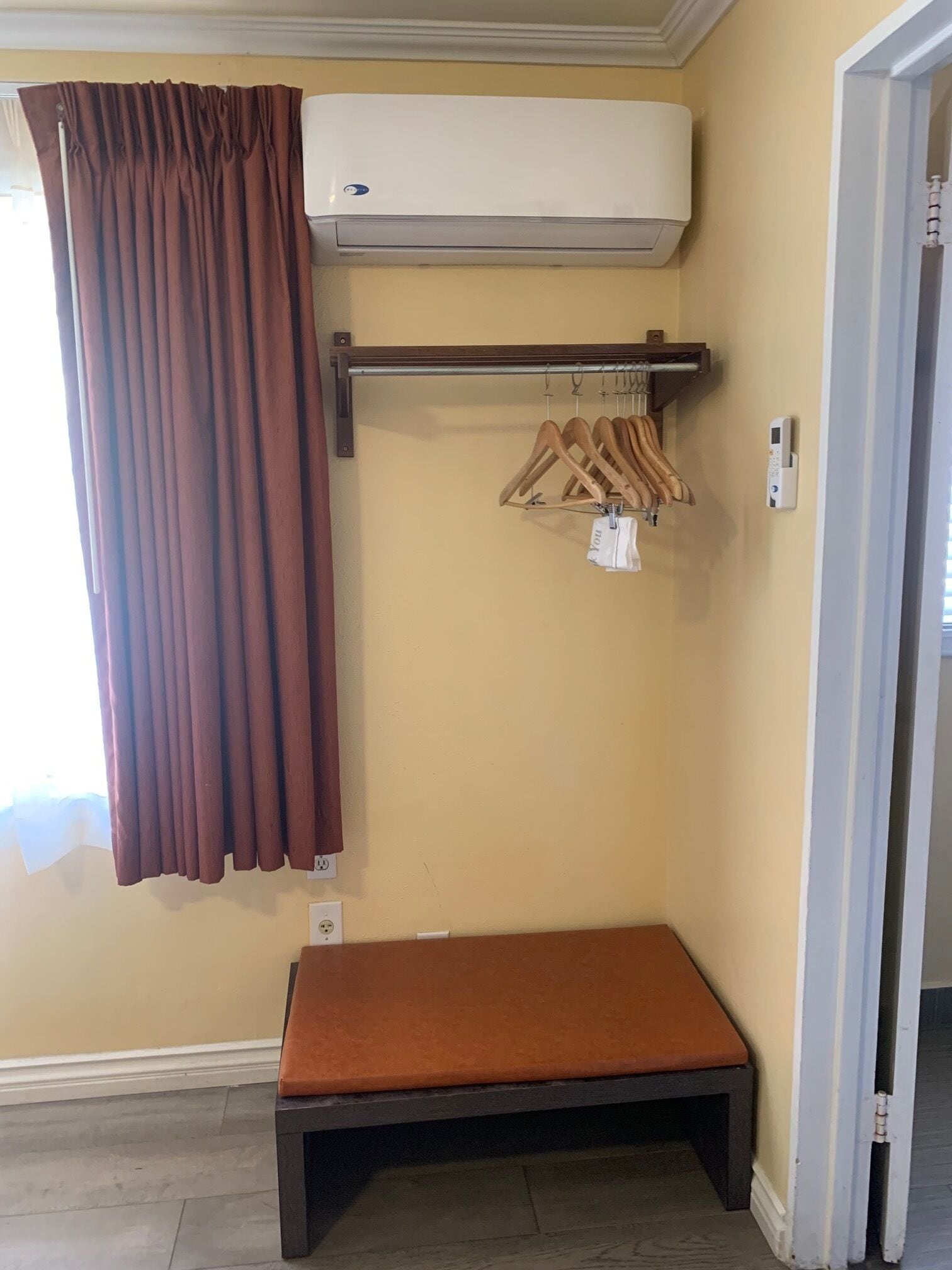A room with a brown curtain, a bench, a wall-mounted air conditioner, and a wooden hanger rack with clothes.