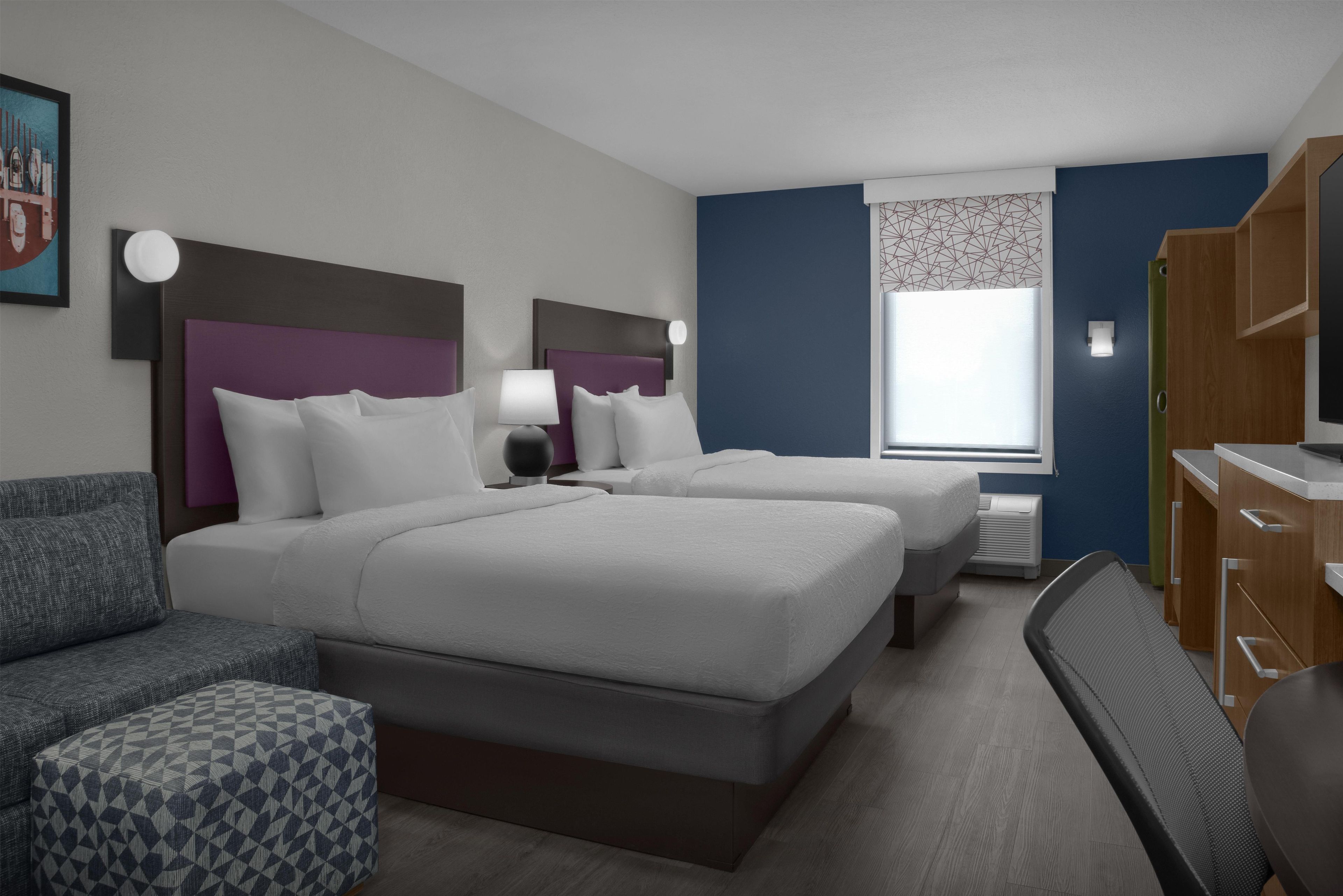 Ein Hotelzimmer mit zwei Betten, einem Schreibtisch, einem Stuhl, einem Sofa und einem gemusterten Hocker.