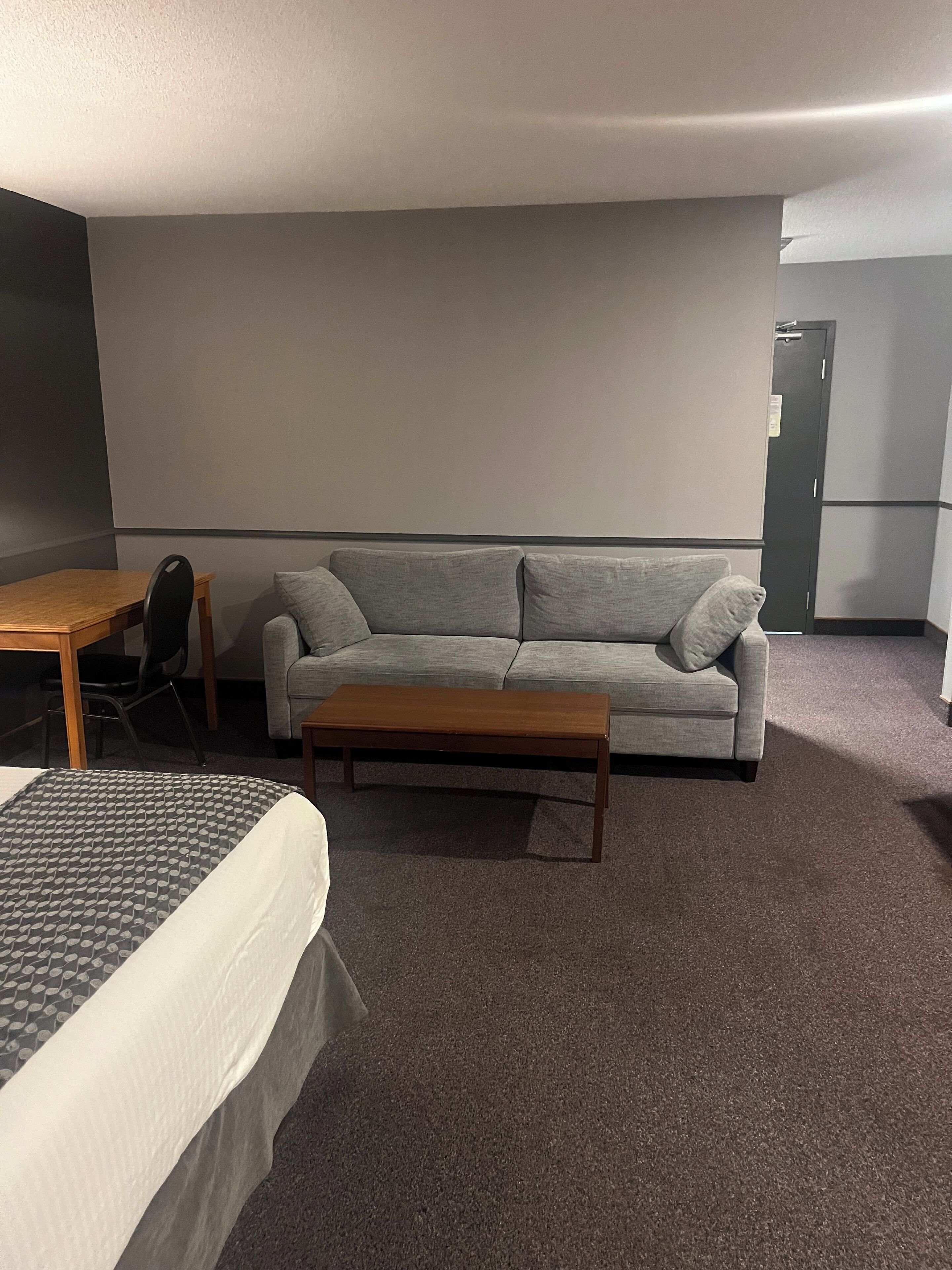 Habitación de hotel con cama, sofá gris, mesita de centro de madera, una pequeña mesa con una silla y una puerta.