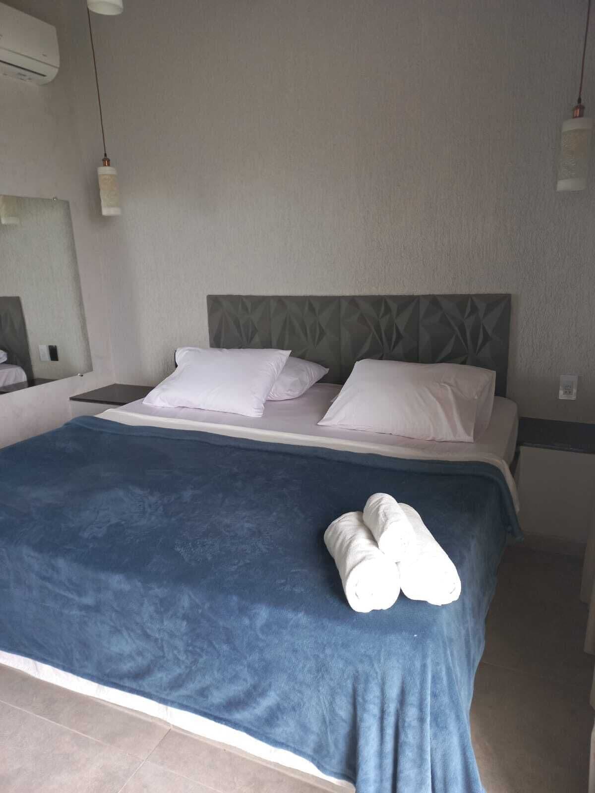 A neatly made bed with a blue blanket, two pillows, and a pair of rolled towels on the side.