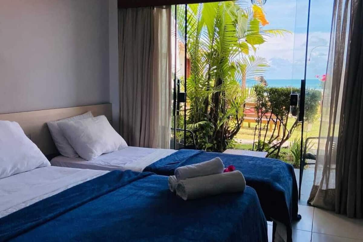 Ein Hotelzimmer mit zwei Betten, einem großen Fenster mit Blick auf den Strand und Palmen sowie einer Deckenleuchte.