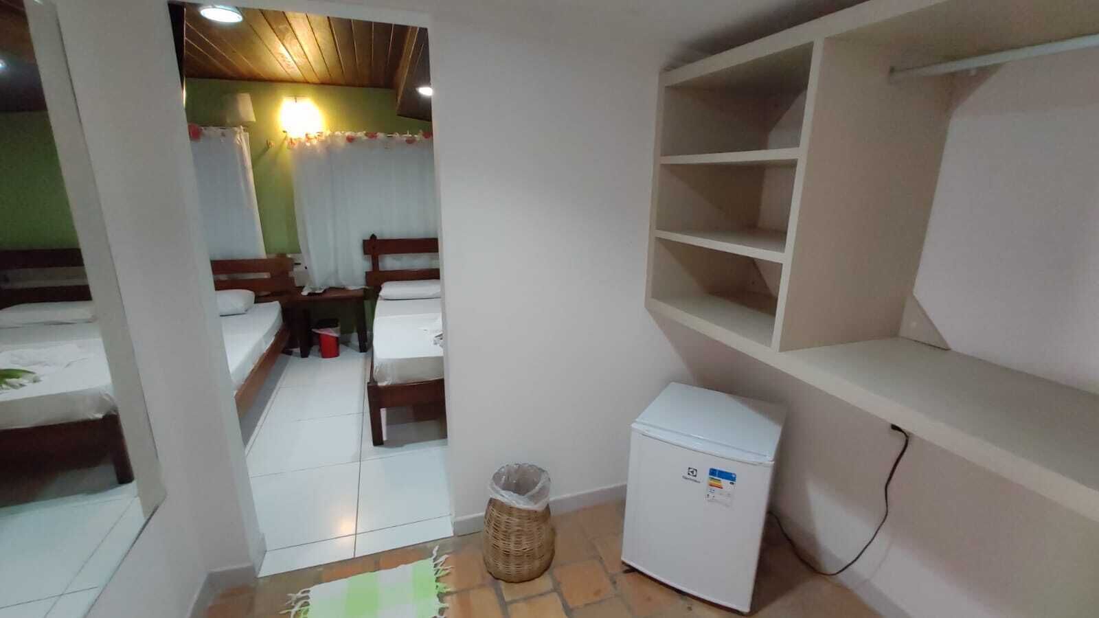 A small, clean room with a bed, a small refrigerator, a trash bin, and a shelf unit.
