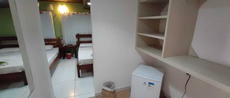 A small, clean room with a bed, a small refrigerator, a trash bin, and a shelf unit.