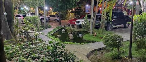 Vista nocturna de un estacionamiento con varios coches, un pequeño estanque y abundante vegetación.