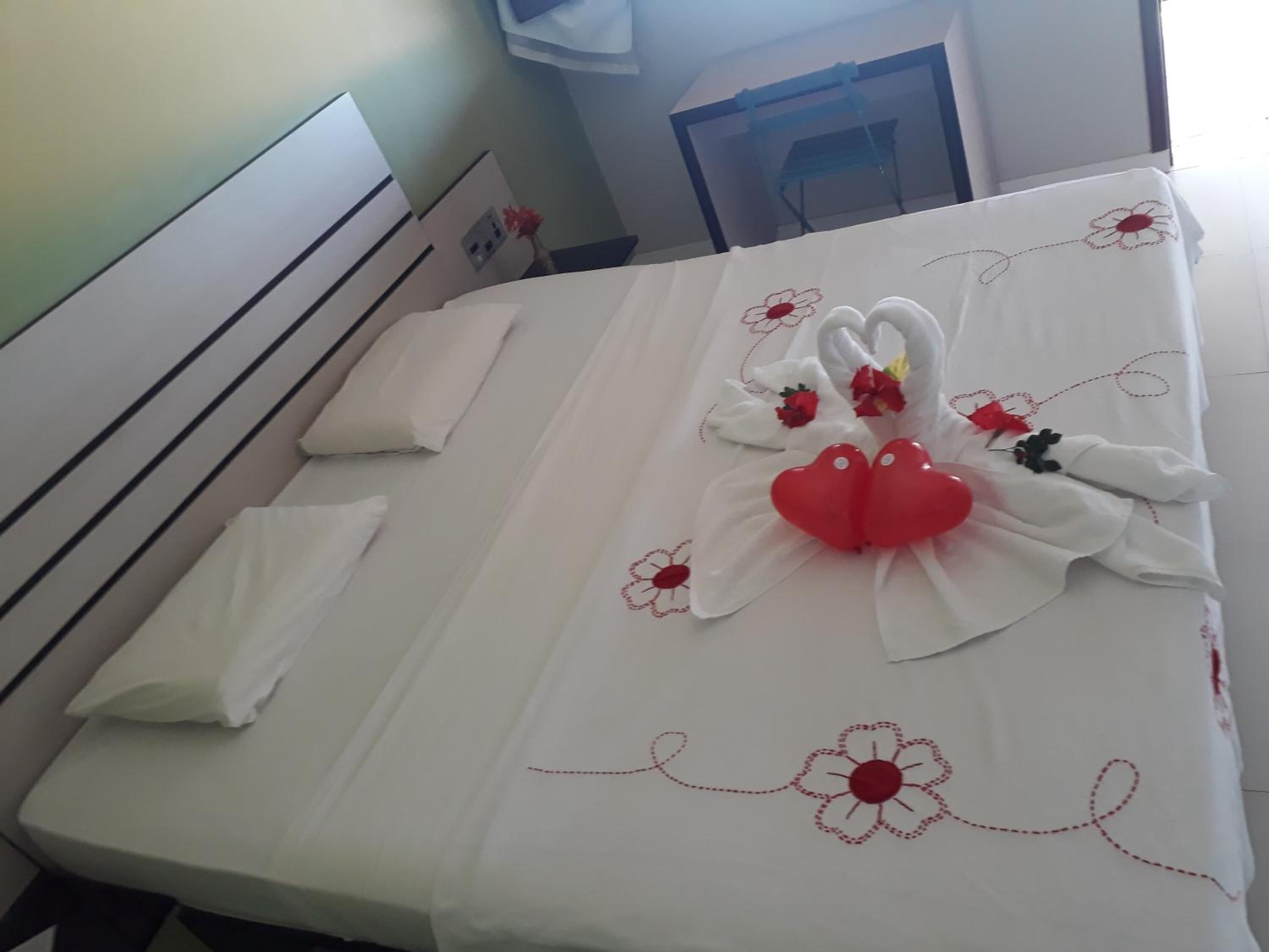 Un lit bien fait, recouvert d’une courtepointe blanche, agrémenté de coussins décoratifs en forme de cygne et de cœur, et surmonté d’un petit bouquet de fleurs.