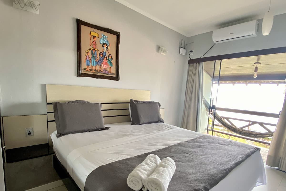 Ein Hotelzimmer mit einem großen Bett, einem Kopfteil, einem gerahmten Bild an der Wand, einer Klimaanlage und einem Fenster mit Blick.