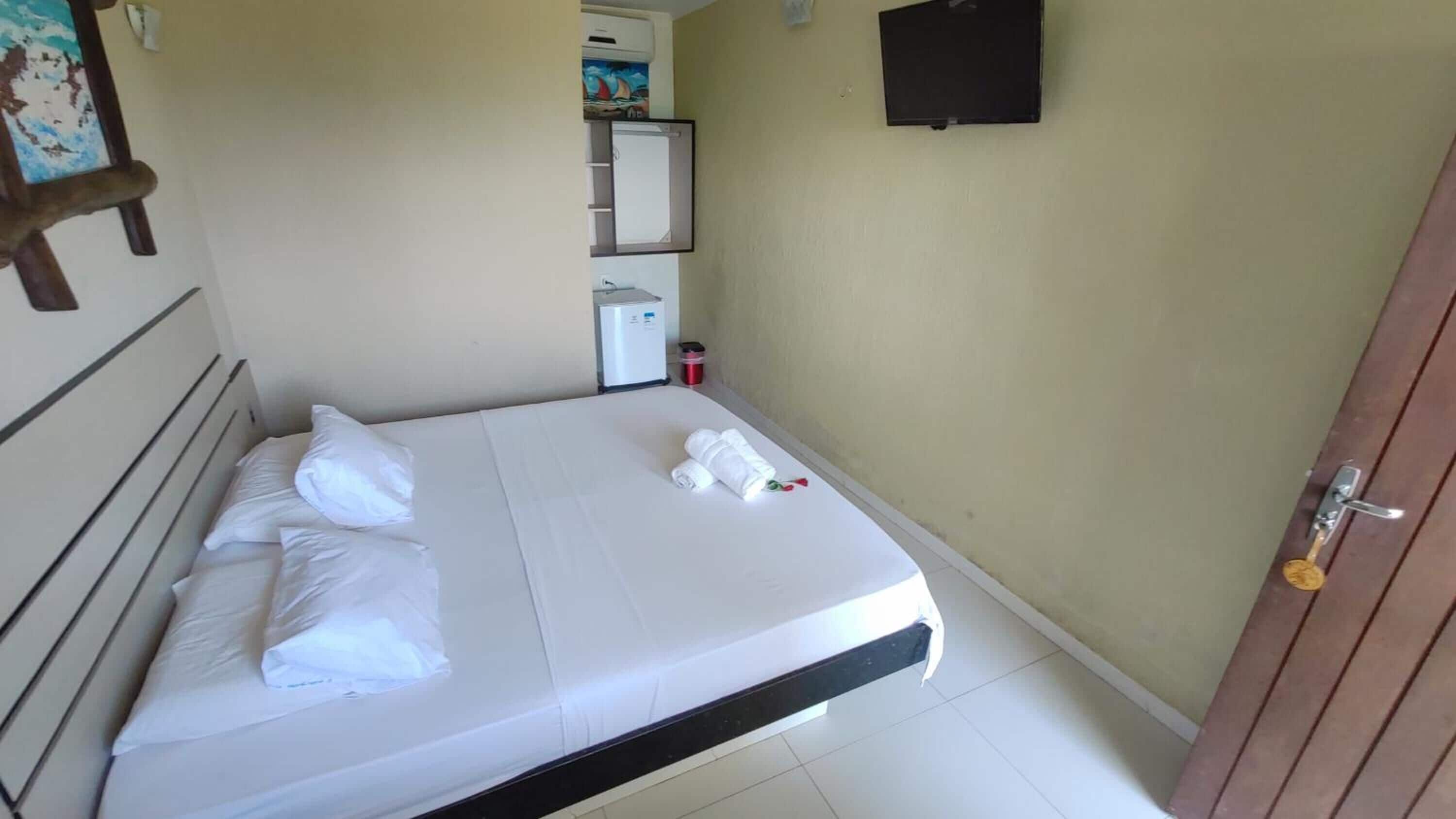 Ein Hotelzimmer mit Bett, Kissen, Handtuch, kleinem Kühlschrank, Mülleimer und Tür.