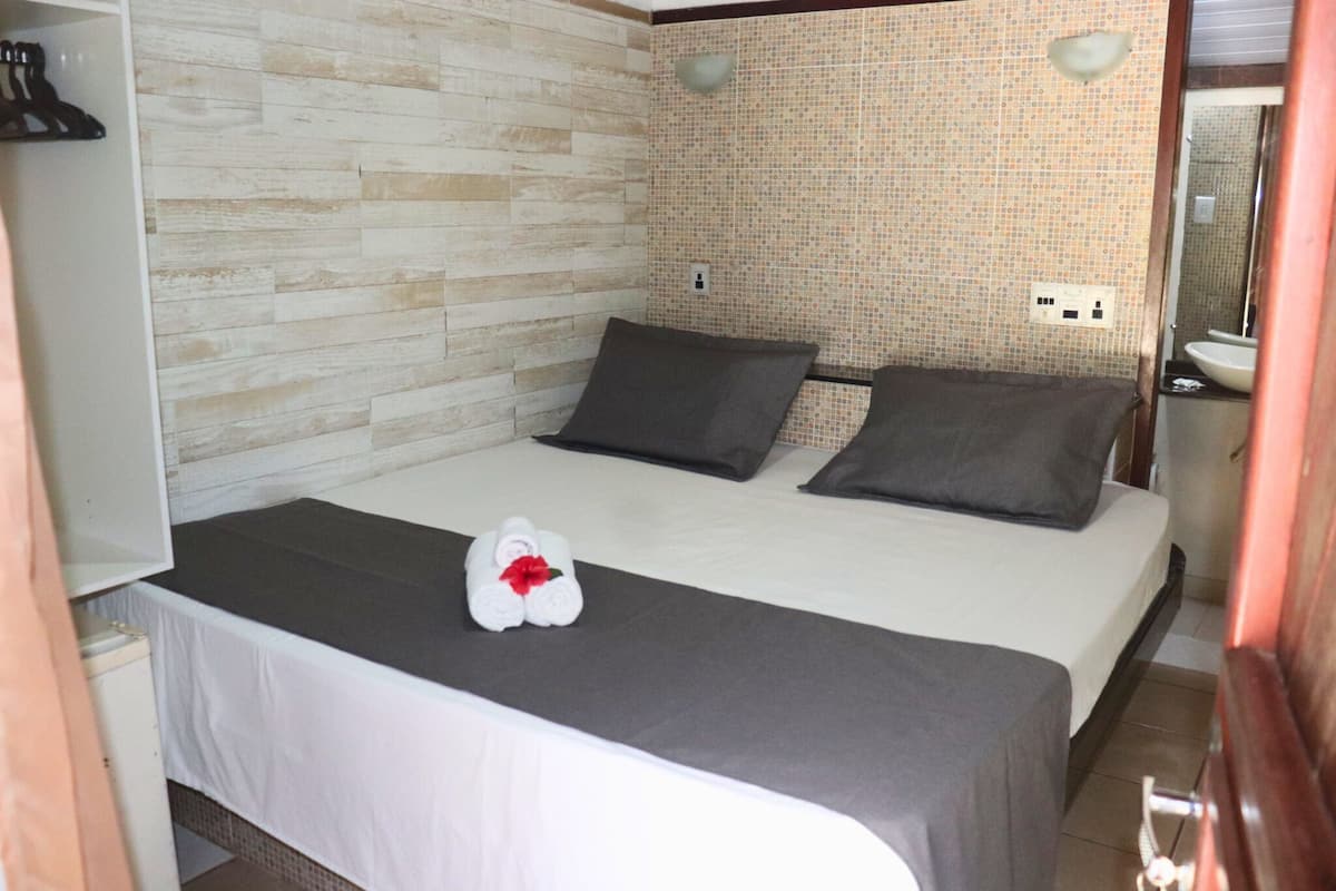 Ein Hotelzimmer mit einem Bett, zwei Kissen, einem weißen Handtuch mit roter Schleife, einer Holzwand, einer Fliesenwand, einem Spiegel und einem Waschbecken.