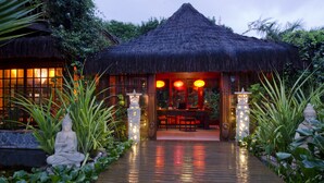 A thatched-roof structure with a wooden deck leading to an indoor seating area, surrounded by lush greenery and a Buddha statue. - Sombra e Agua Fresca Floresta (Tibau do Sul)