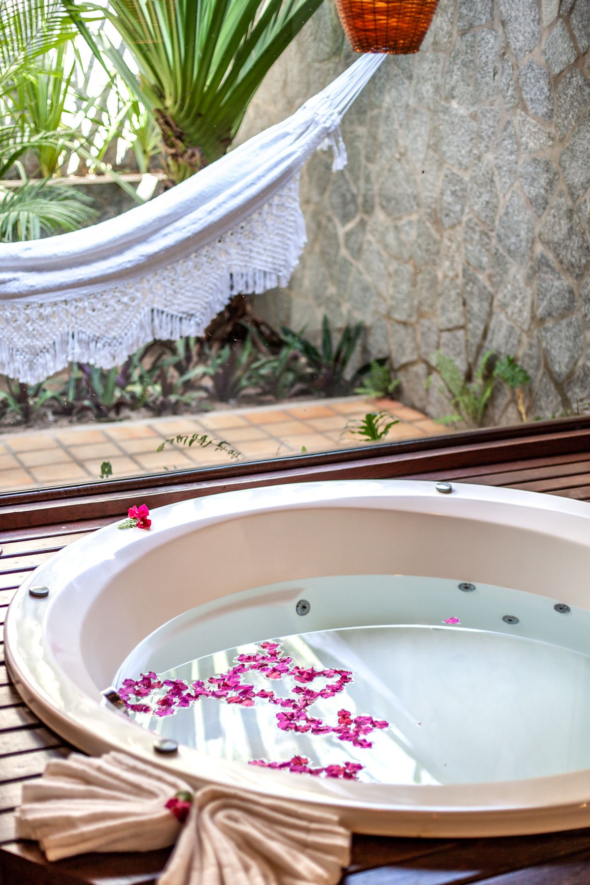 A hot tub with pink flower petals floating on the water, surrounded by a hammock and potted plants.