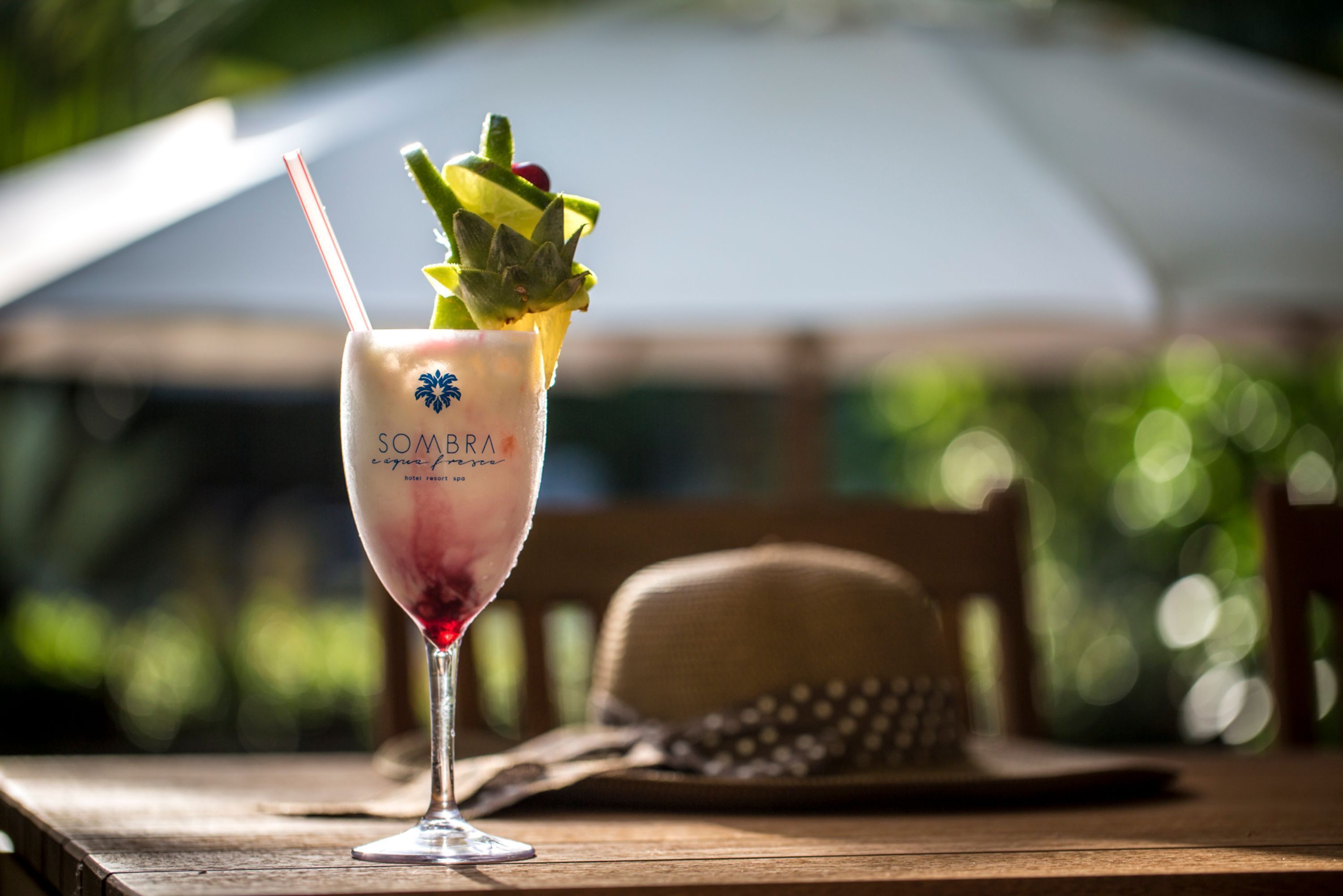 A glass of cocktail with a straw and garnishes, placed on a wooden table with a hat and umbrella in the background.