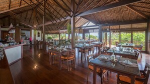 A spacious restaurant with wooden tables and chairs, a thatched roof, and large windows offering views of greenery. - Sombra e Agua Fresca Floresta (Tibau do Sul)