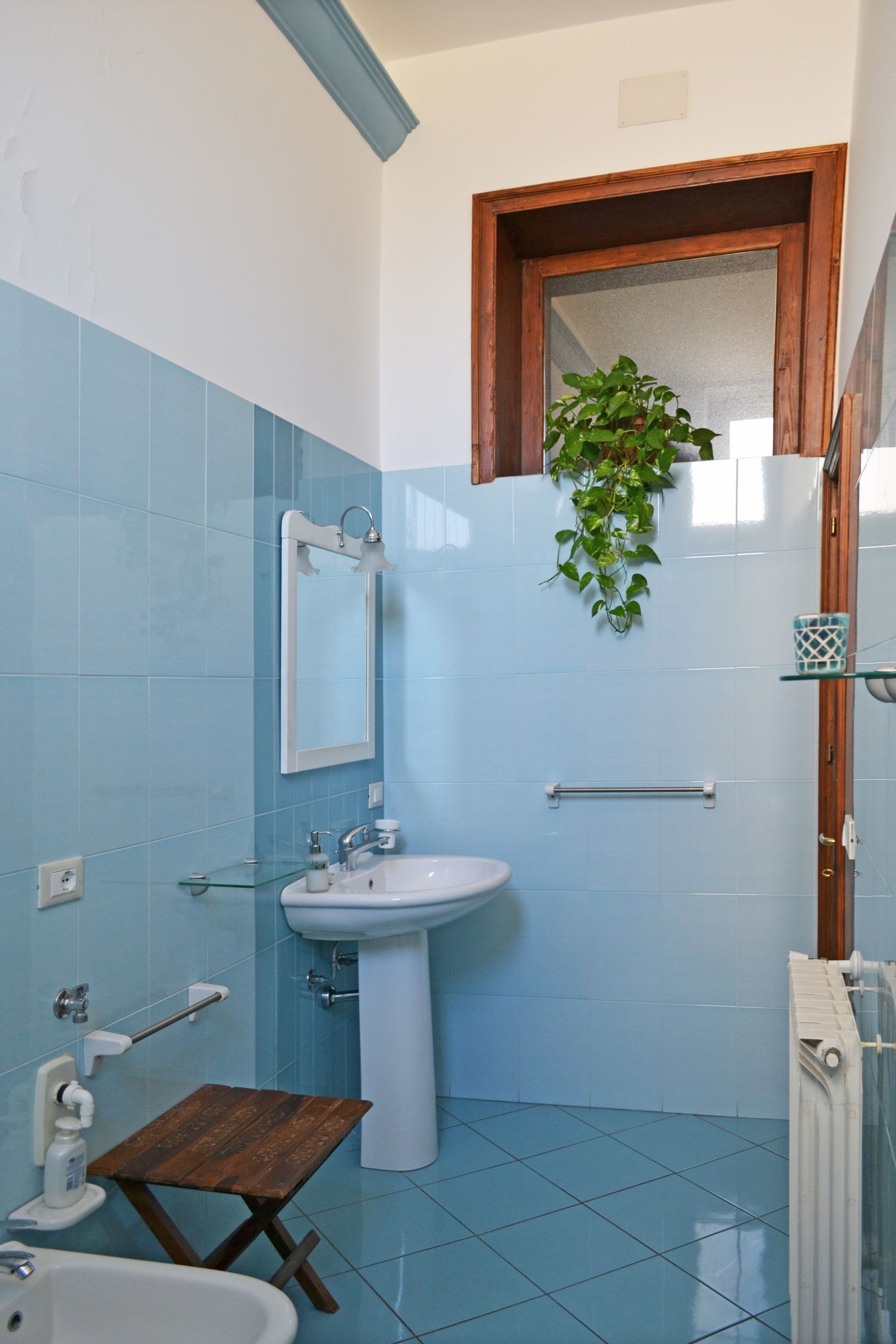 A bathroom with blue tiles, a white sink, a mirror, a wooden stool, and a hanging plant.