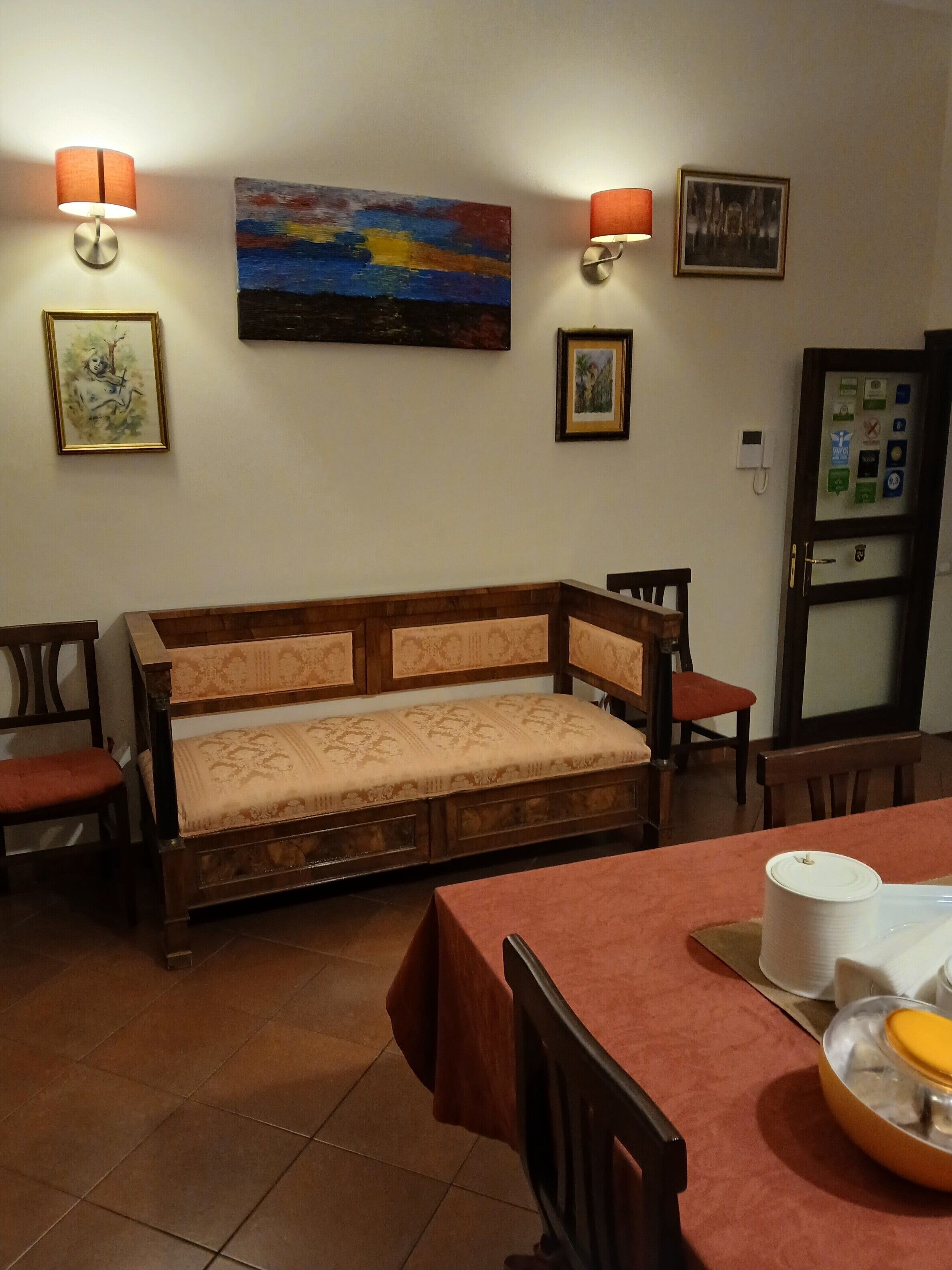 Une pièce chaleureuse avec un canapé en bois, une table à manger avec des chaises et un réfrigérateur. Des lampes murales et des tableaux encadrés ornent les murs.