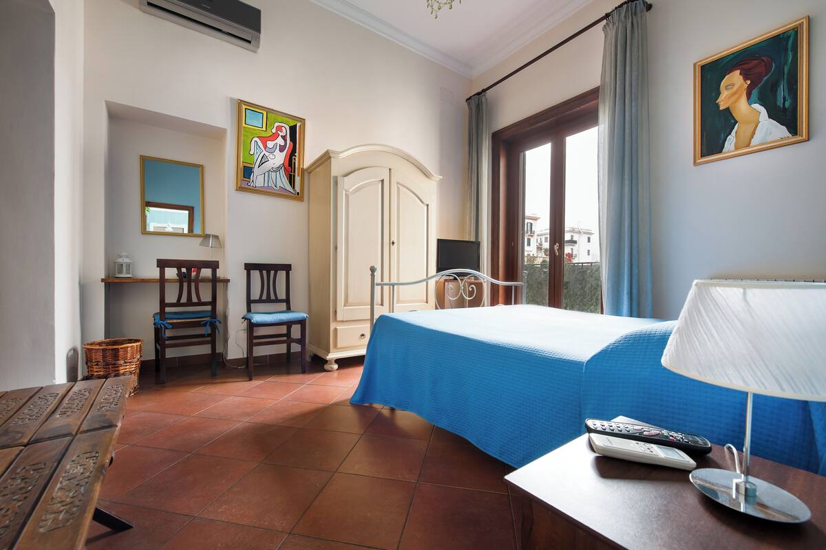 Habitació d'hotel amb llit, escriptori, cadira, làmpada i finestra amb cortines.